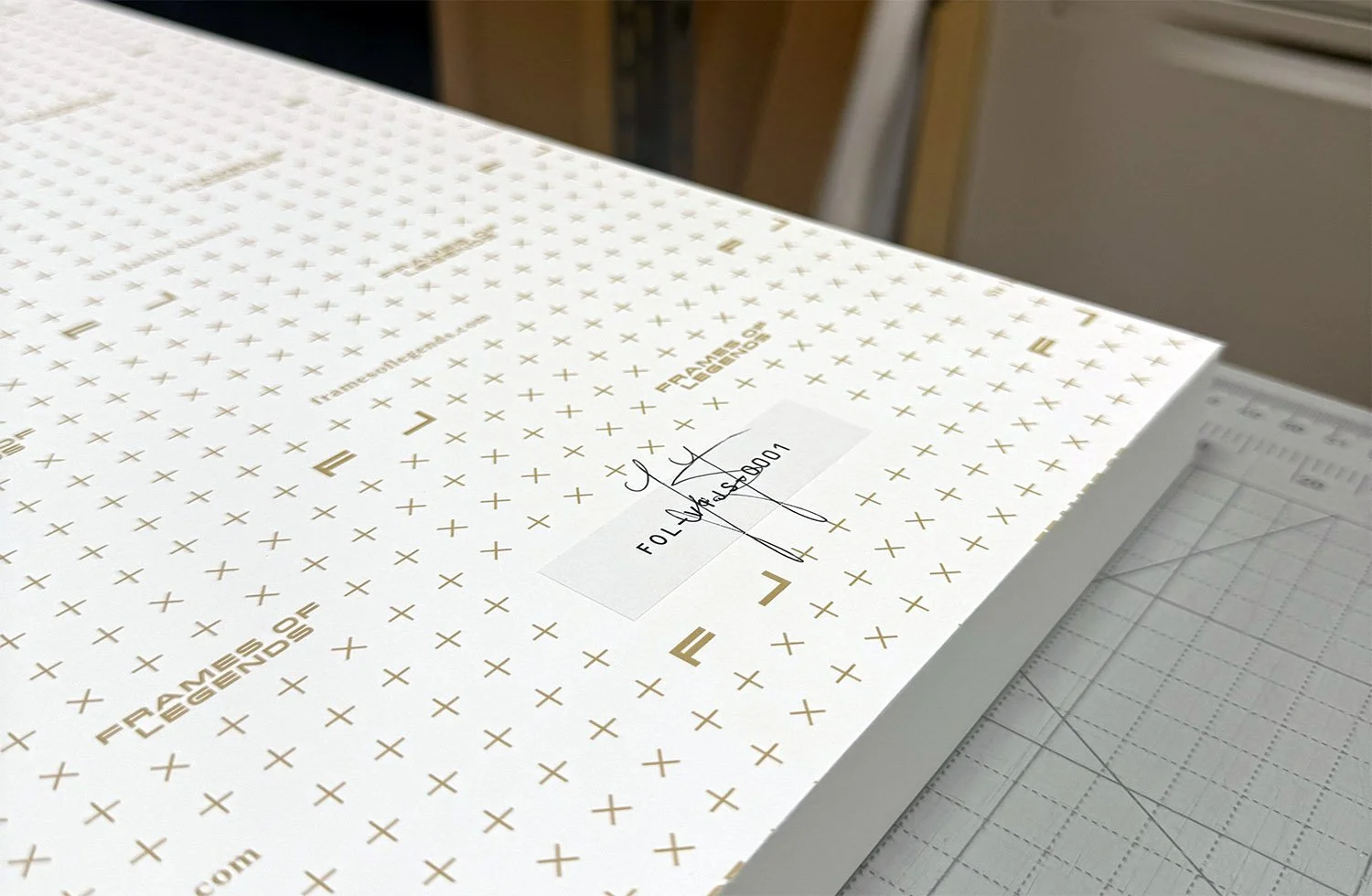 A large sheet of white foam core with gold lettering and crosses, marked 'FRAMES & LEGENDS' and a label with a signature and code, resting on a cutting mat in a workspace.