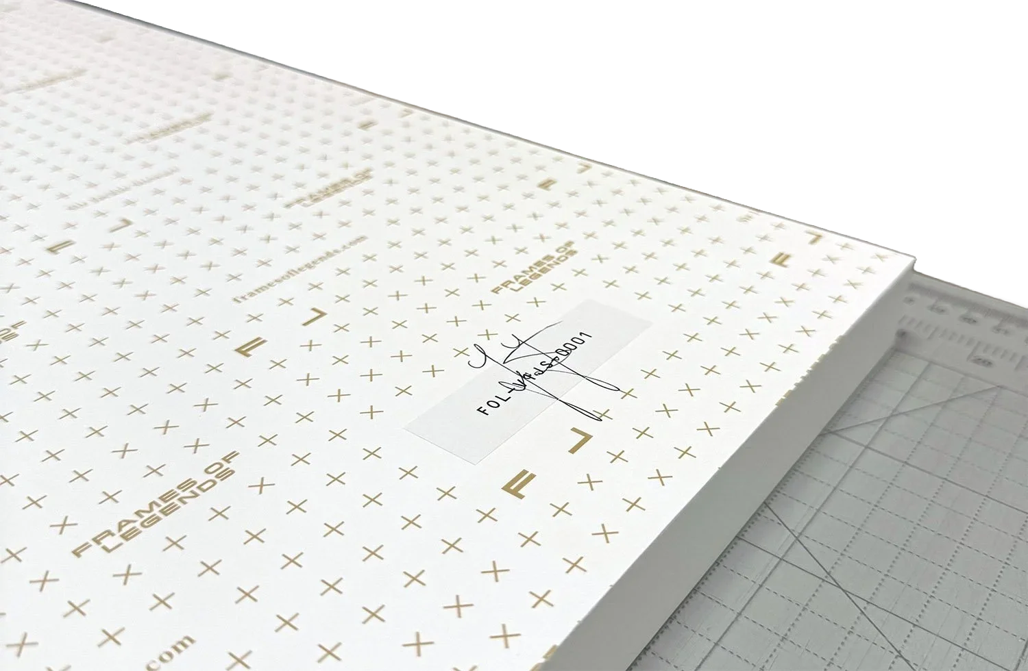 A large sheet of white foam core with gold lettering and crosses, marked 'FRAMES & LEGENDS' and a label with a signature and code, resting on a cutting mat in a workspace.