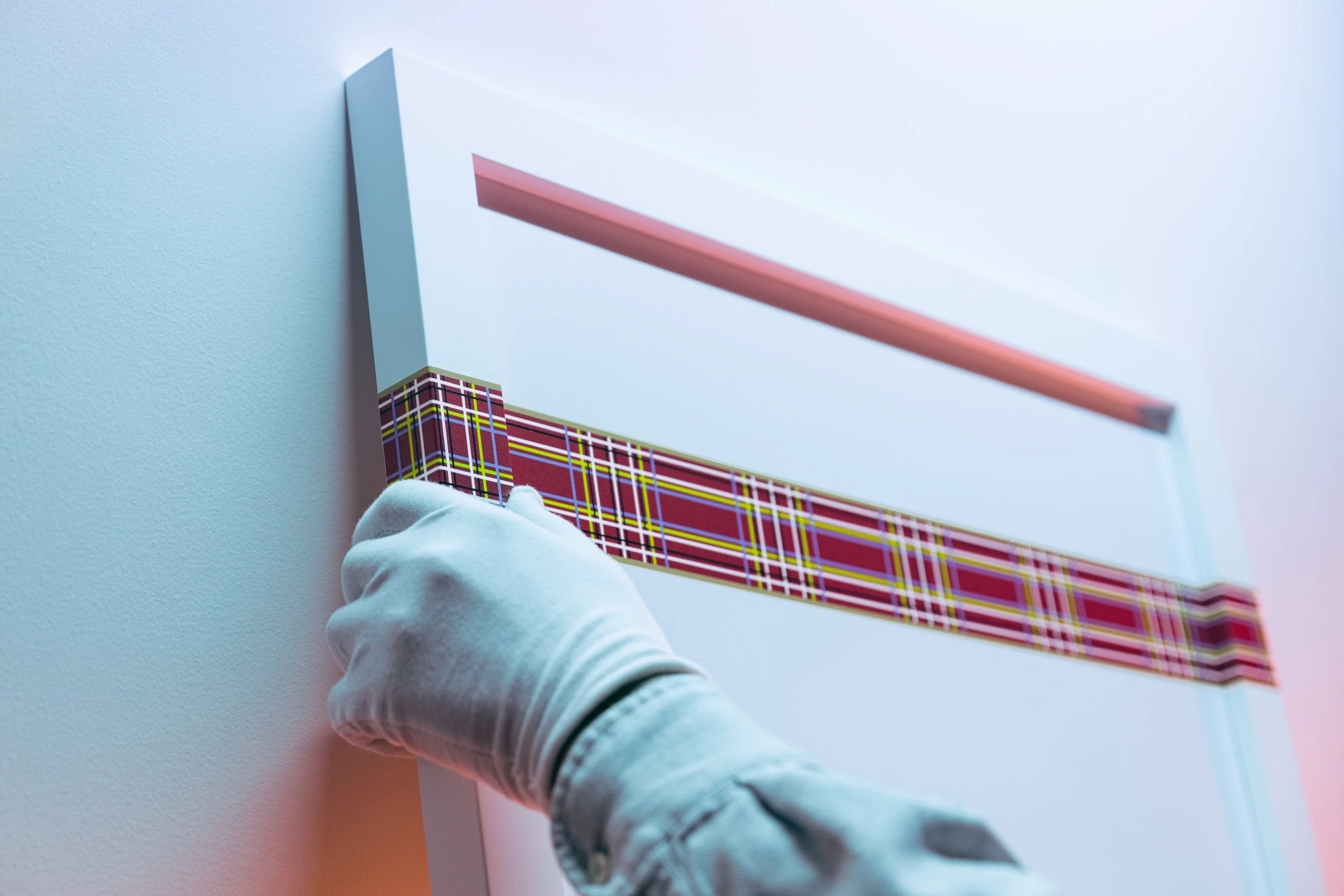 A gloved hand holding a rectangular box with a red, yellow, green, and white plaid pattern, against a light blue wall and white background.