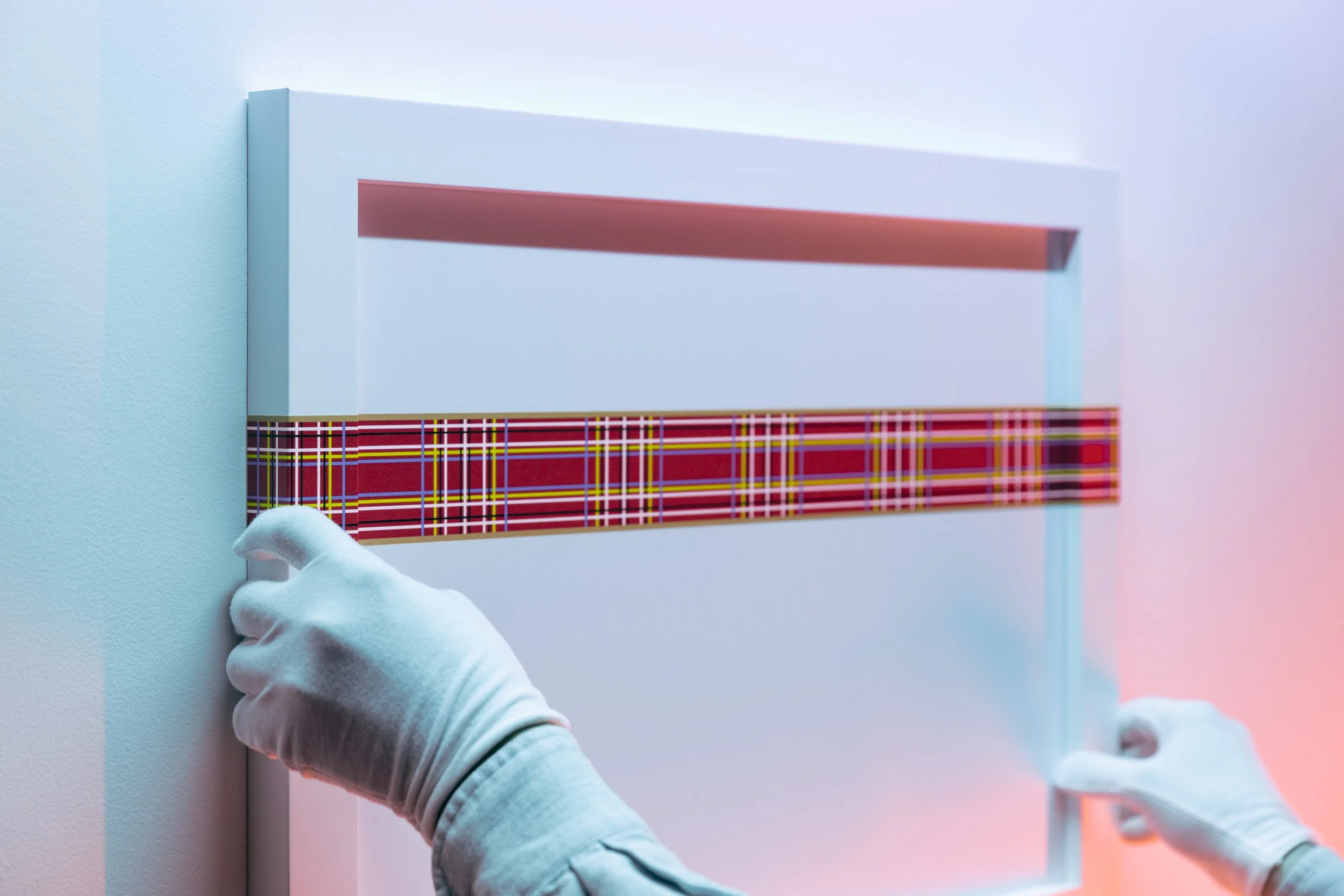A person wearing white gloves is installing or adjusting a white panel with a red stripe and a colorful tartan pattern at the bottom, on a white wall.