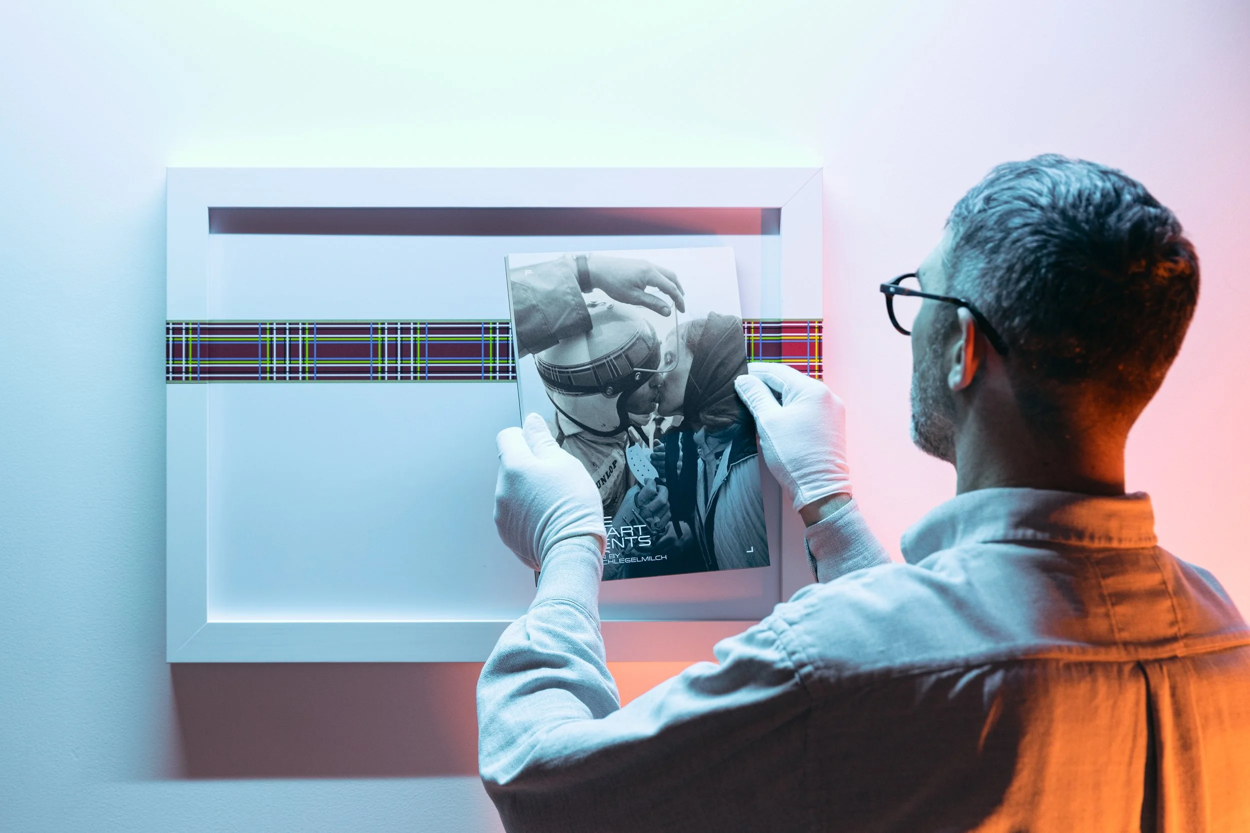 A man wearing glasses and gloves is holding a black-and-white photograph of a person in a helmet, with a colorful strip decoration behind them.