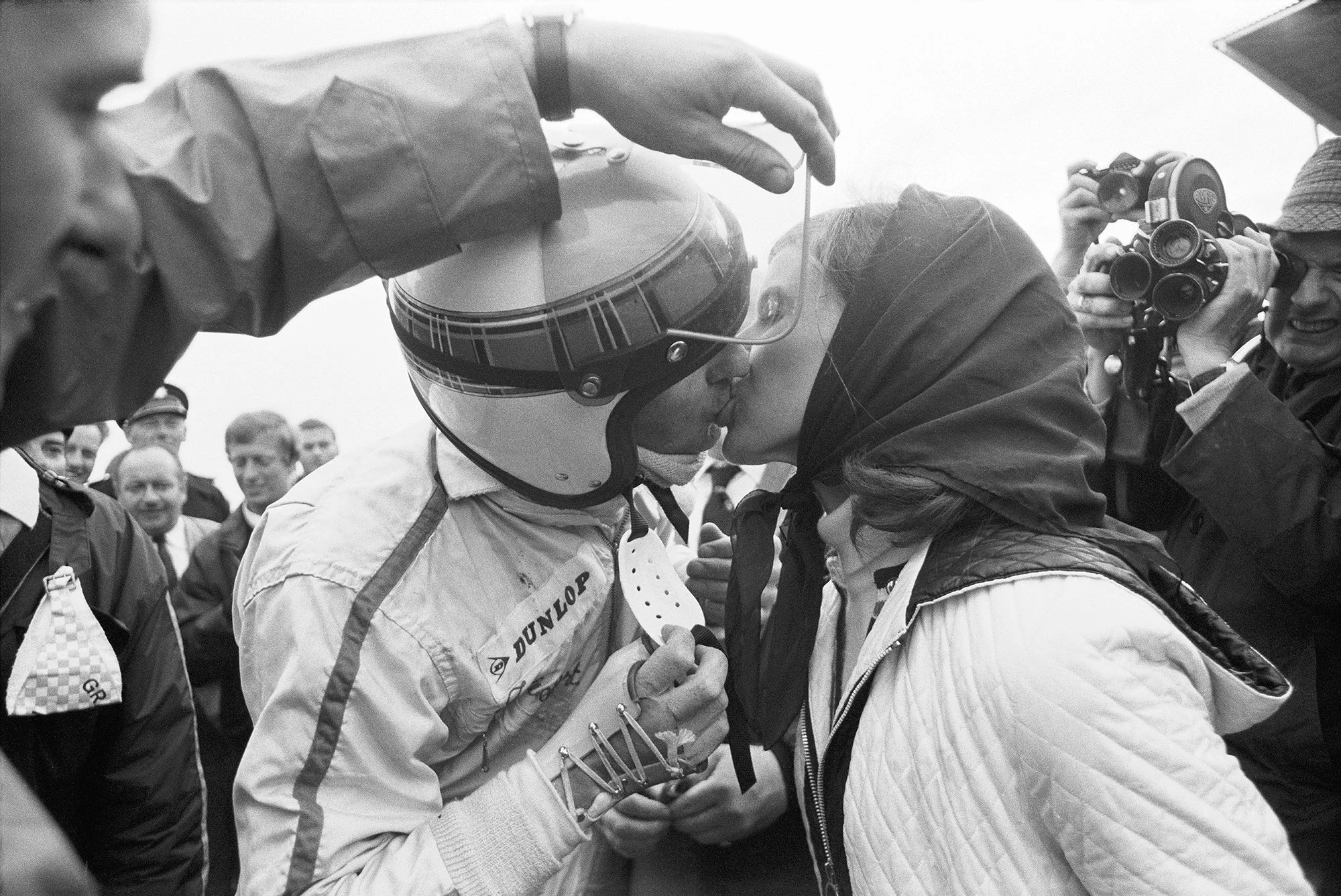 Two race car drivers in racing suits sharing a pen, with one wearing a helmet and the other with glasses under her helmet, surrounded by spectators and photographers, in black and white.