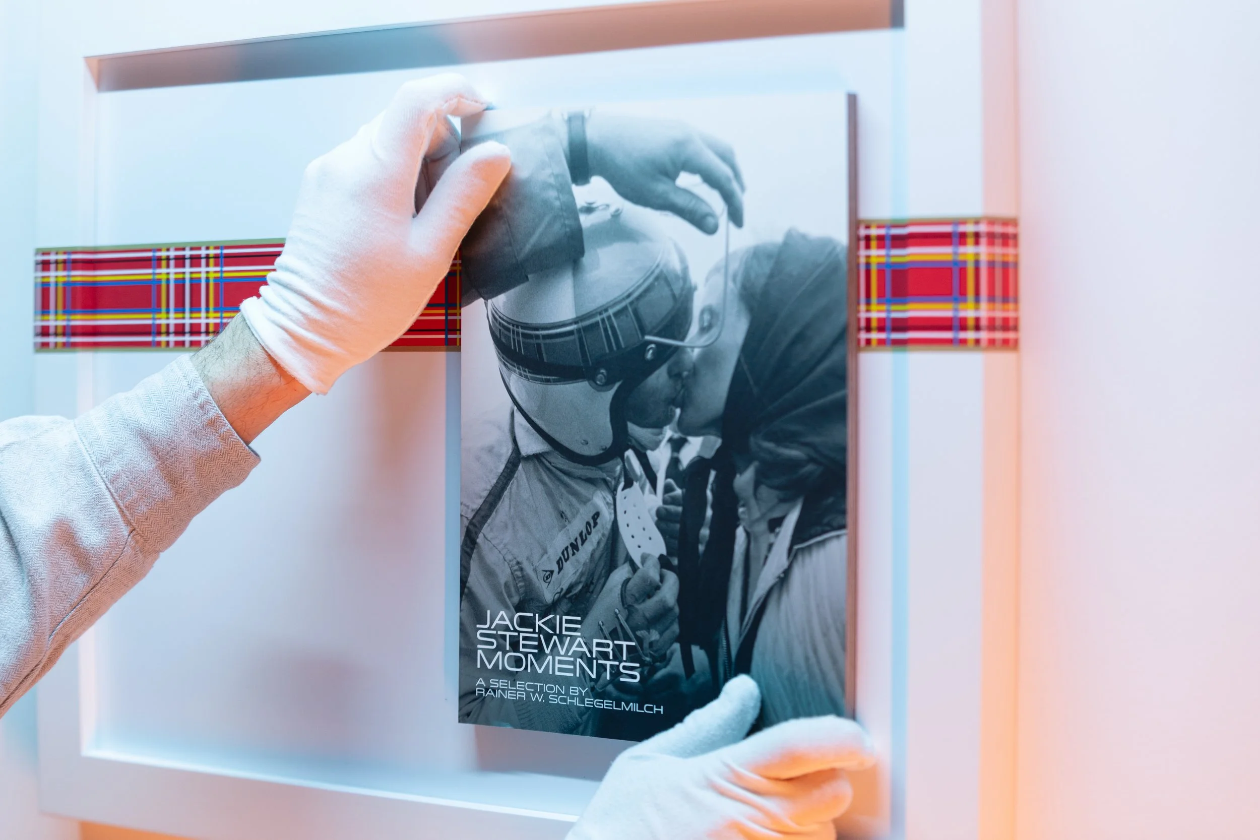 A person wearing white gloves holding a framed black-and-white photograph of two people, one in racing gear and helmet, and the other in a cap, with text on the photograph indicating it is a collection of Jackie Stewart moments.