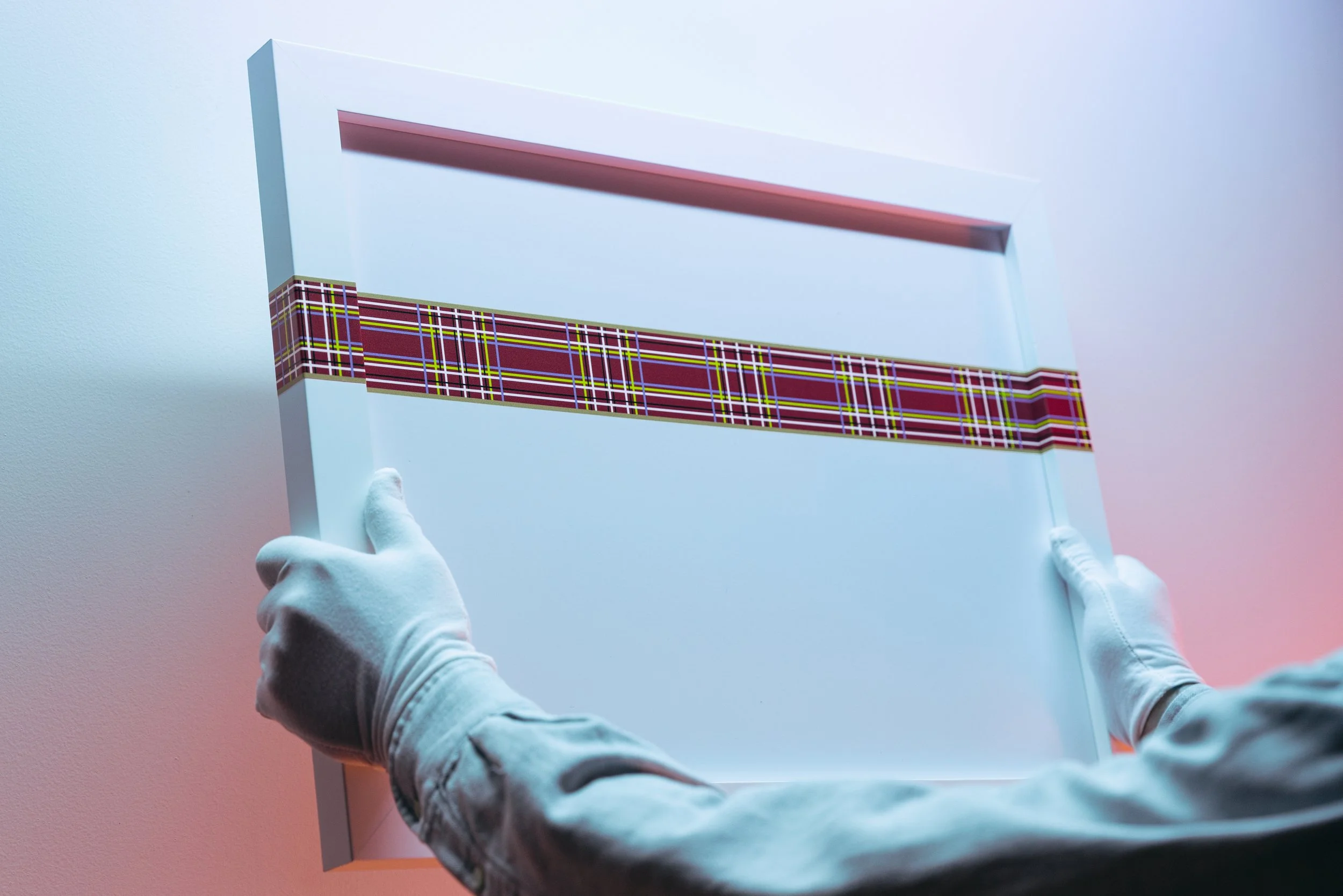 Person wearing white gloves holding a rectangular mirror with a red and black striped pattern on the frame, against a light blue background.