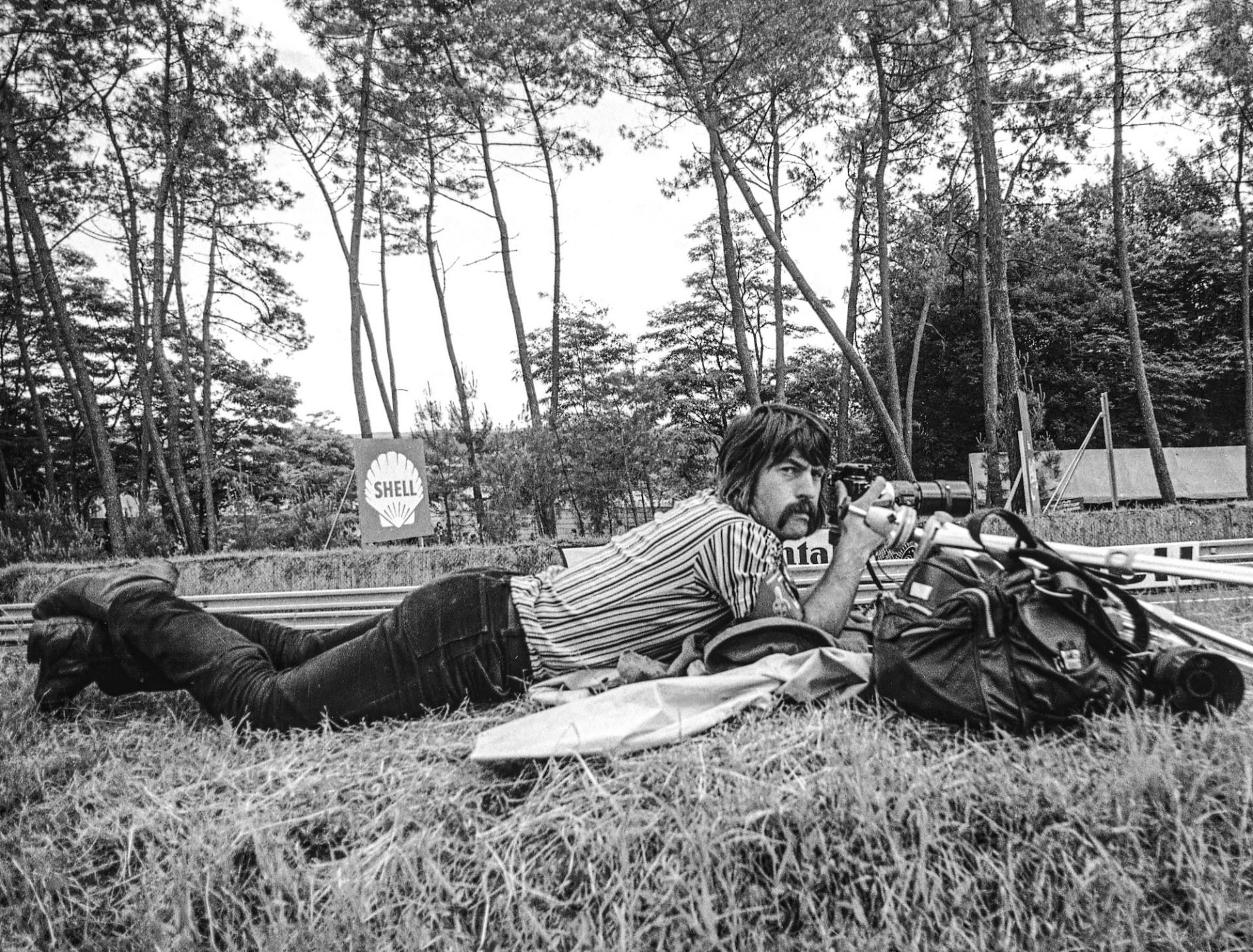 A person lying on the ground outdoors in a wooded area, aiming a rifle with a scope. The person has dark hair and a beard, wearing a striped shirt and dark pants. A Shell sign and a Roblox sign are visible in the background.