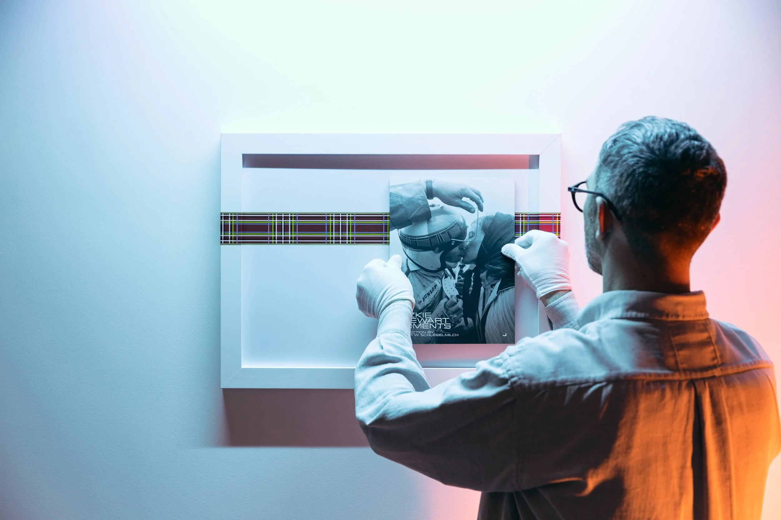 A man with dark hair and glasses, wearing a gray shirt, is looking at a framed black-and-white poster of two racing drivers in helmets and racing gear, with a colorful plaid strip across the middle of the poster. The man is adjusting or hanging the poster on a plain wall.