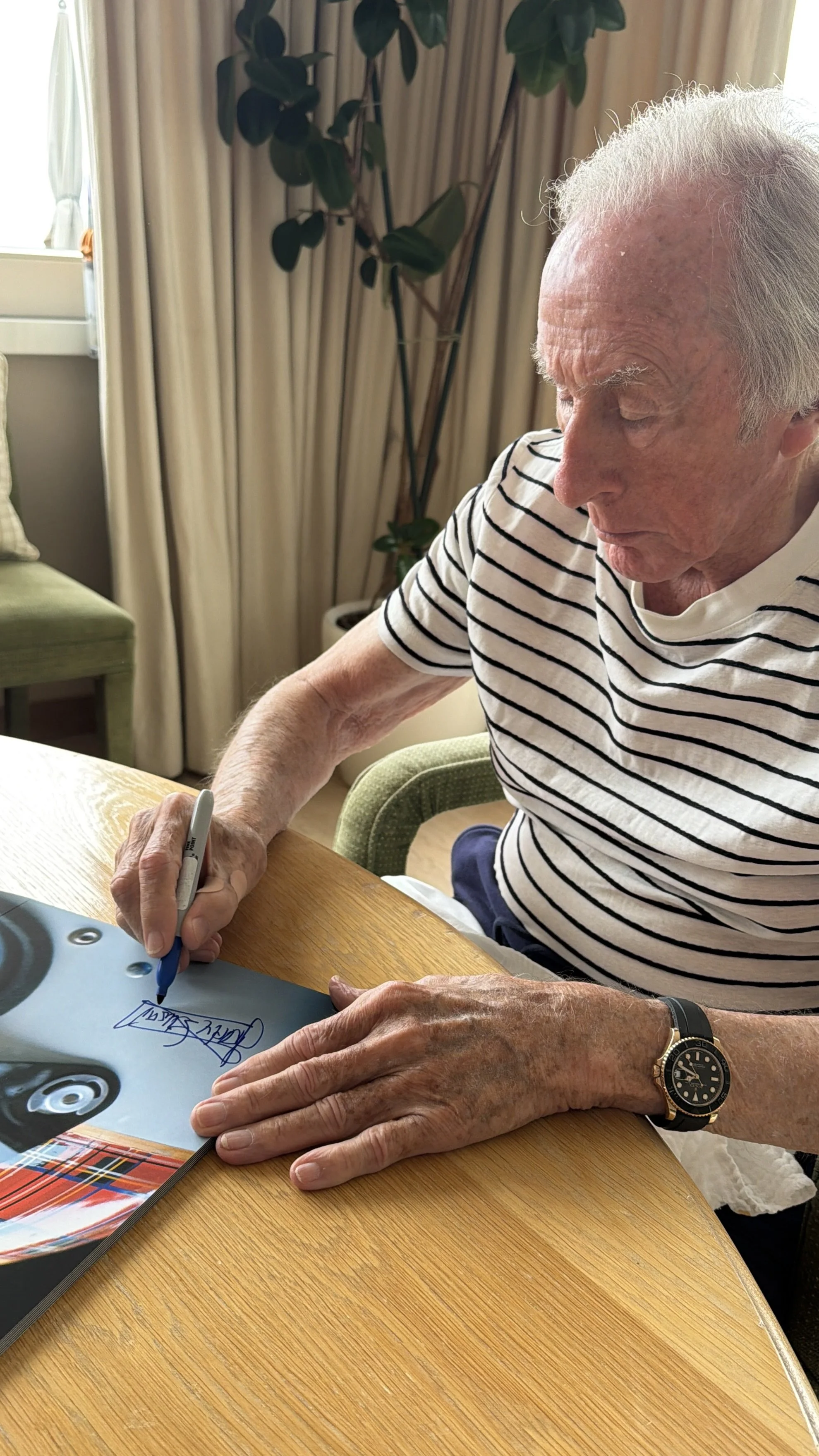 An elderly man with white hair wearing a striped shirt is sitting at a wooden table, signing a picture with a blue marker. There is a large houseplant and curtains in the background.