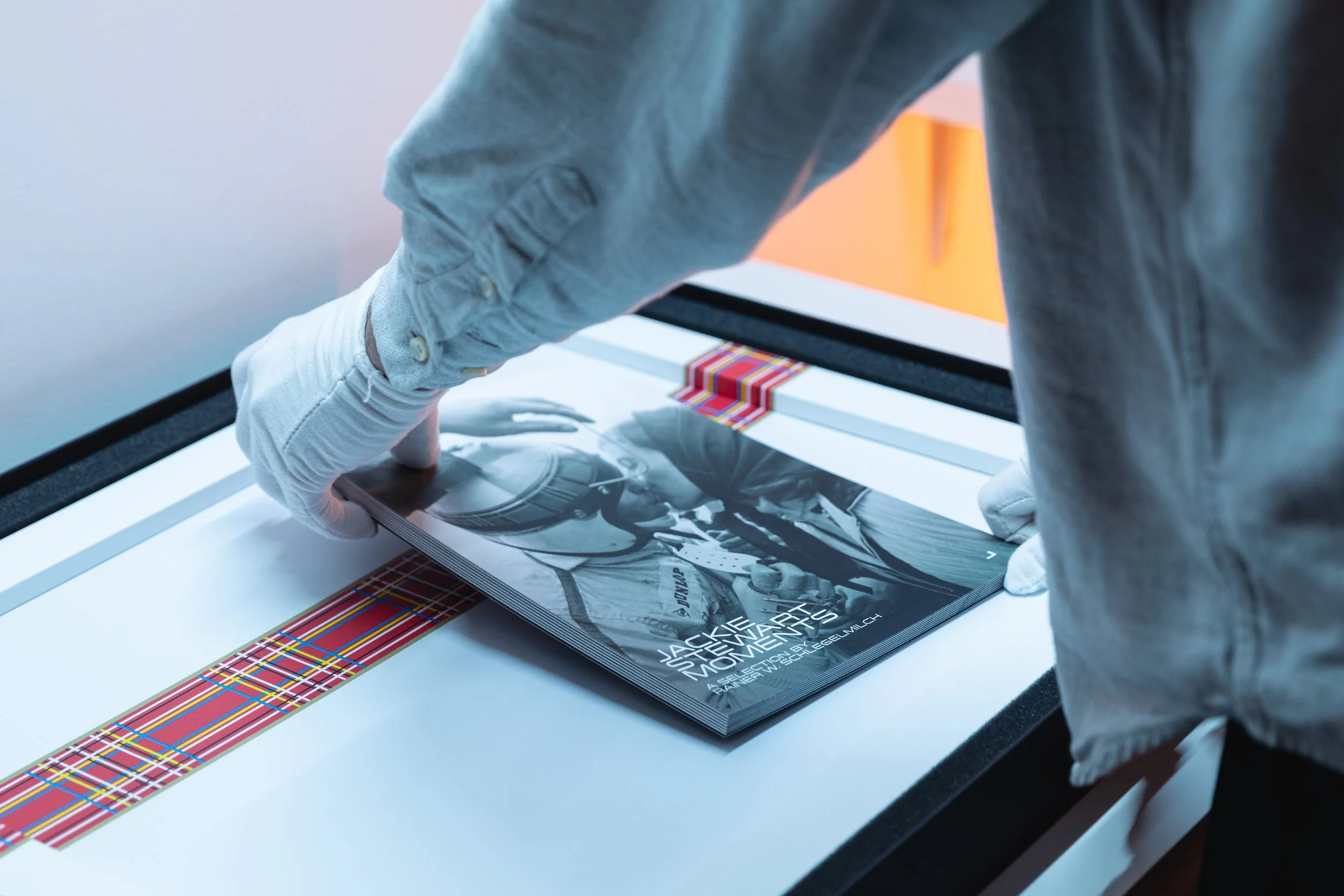 Close-up of a person with gloves handling a magazine titled "Jackie Stewart 50 Years of Moments" on a white table with a red, yellow, and blue plaid pattern along the edge.