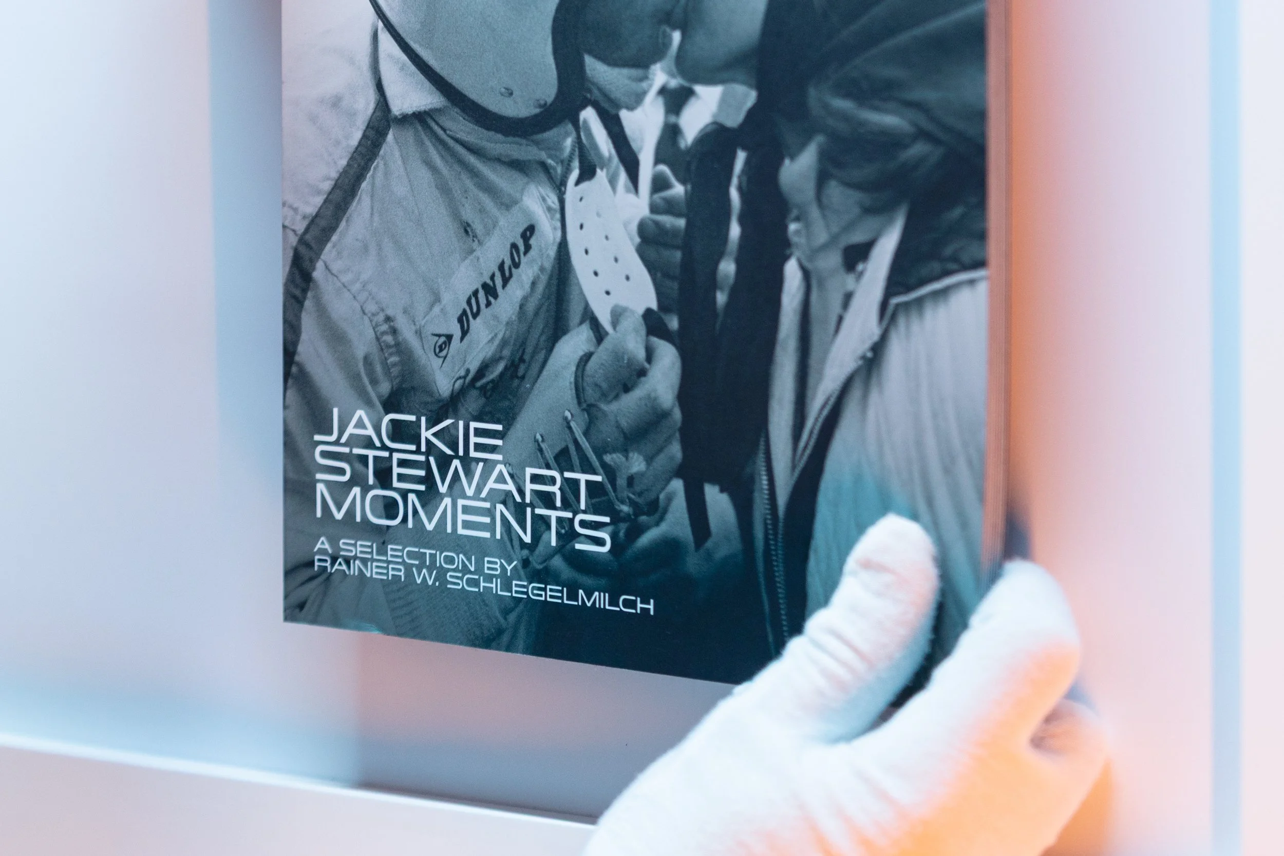 Close-up of a poster featuring a black and white photo of two people with helmets and racing jackets, with text reading "Jackie Stewart Moments: A Selection by Rainer W. Schlegelmilch." A person wearing white gloves is holding the poster.