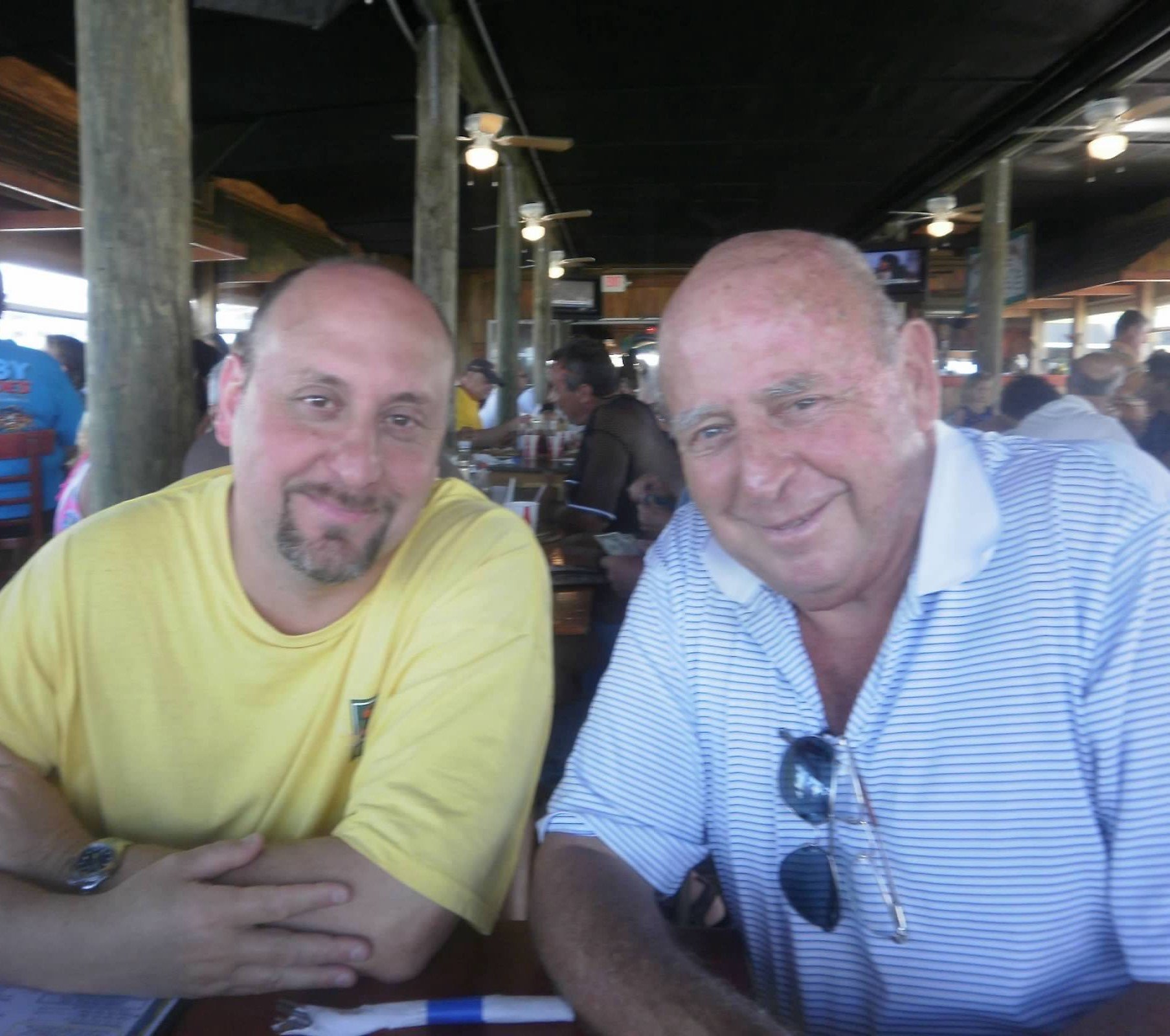 Two men sitting at a restaurant table, smiling at the camera, with a busy restaurant background.