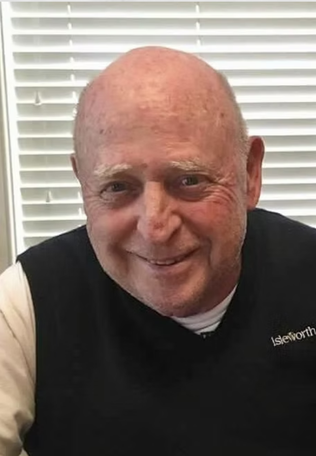 Wally, a bald man with light skin and a beard, wearing a black vest over a light-colored shirt, in front of white window blinds.