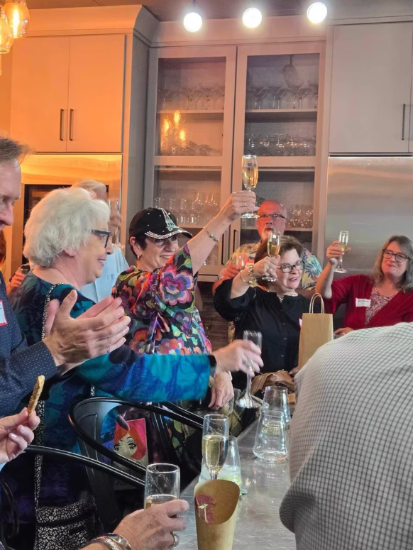 People at a celebration raising glasses of champagne in a kitchen with gray cabinets, glassware, and warm lighting.