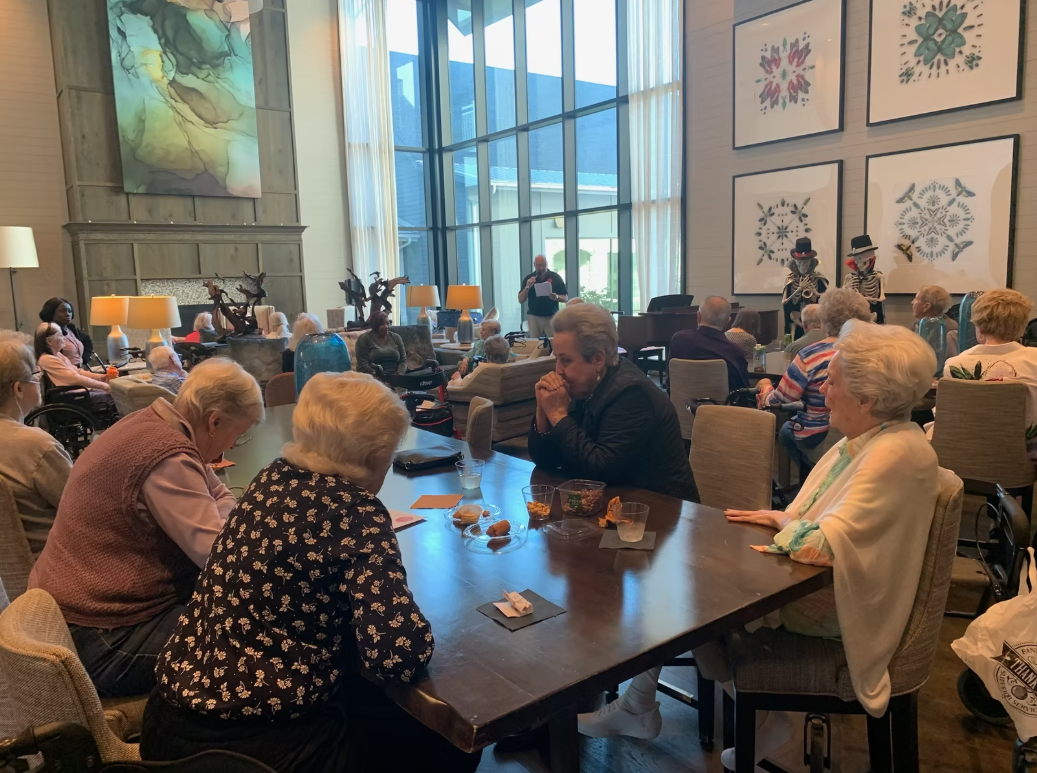 A group of elderly people gathered in a spacious, well-lit room watching a performance. Two performers dressed in skeleton costumes with hats and sunglasses are entertaining the audience. The room has large windows, wall art, and multiple lamps, creating a cozy atmosphere.
