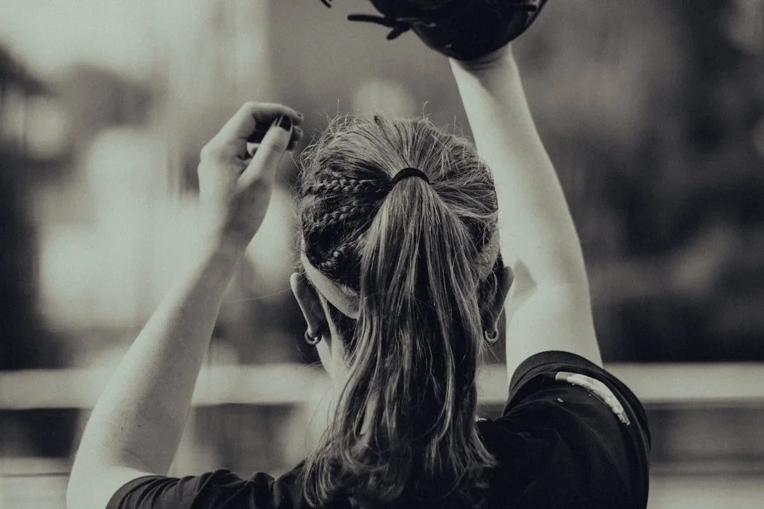 Rear view of a woman with braided and ponytail hair, wearing earrings, raising her hand, and holding a black object in front of a window.