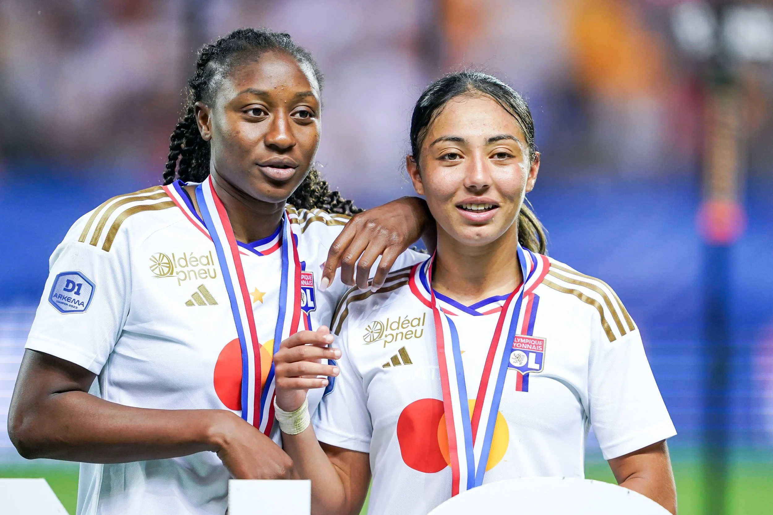Two Olympique Lyonnais Féminin players wearing medals and smiling after winning a championship.