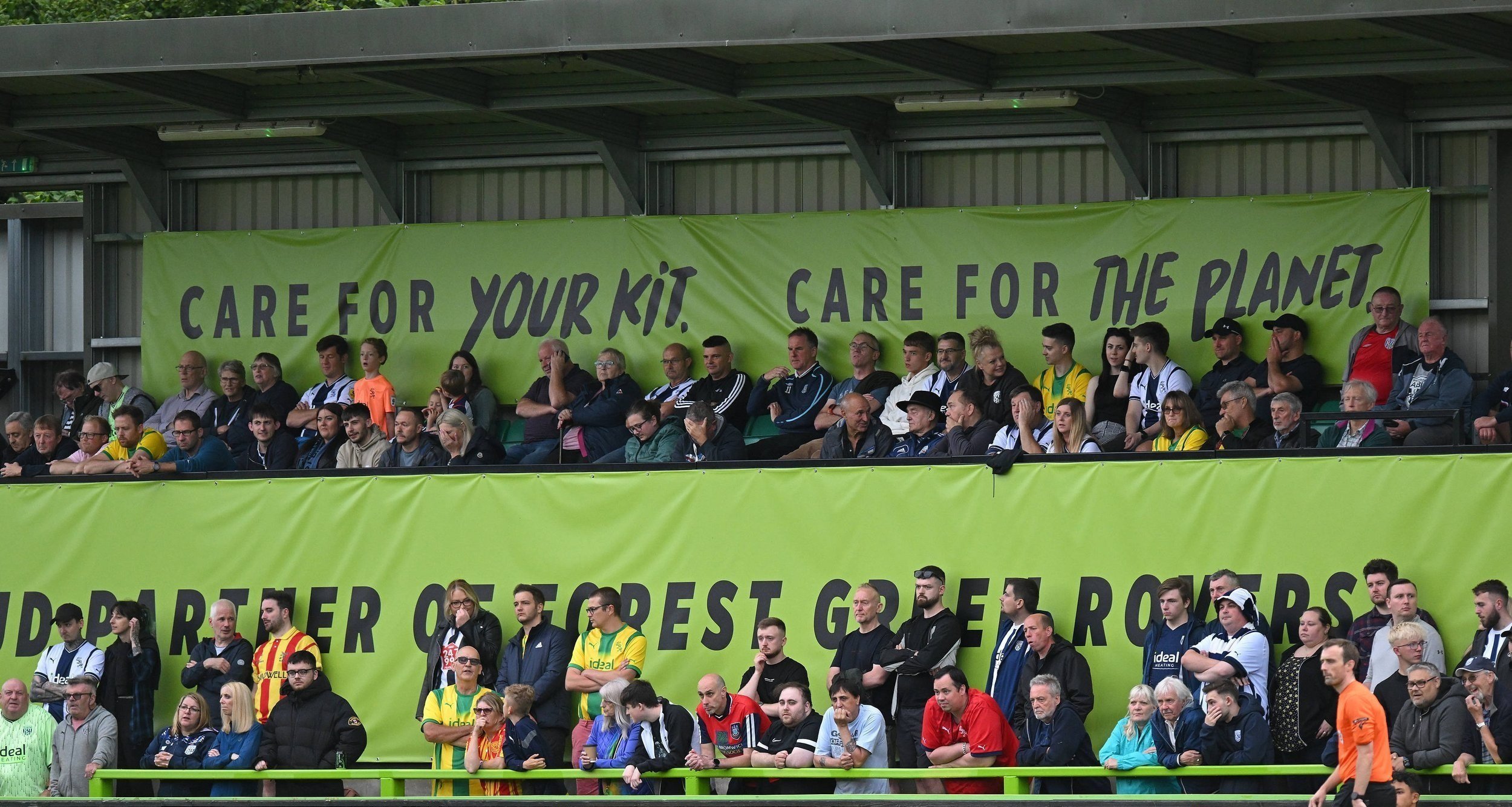 Fans at a Forest Green Rovers stadium next to a sign reading “Care for your kit, care for the planet.”