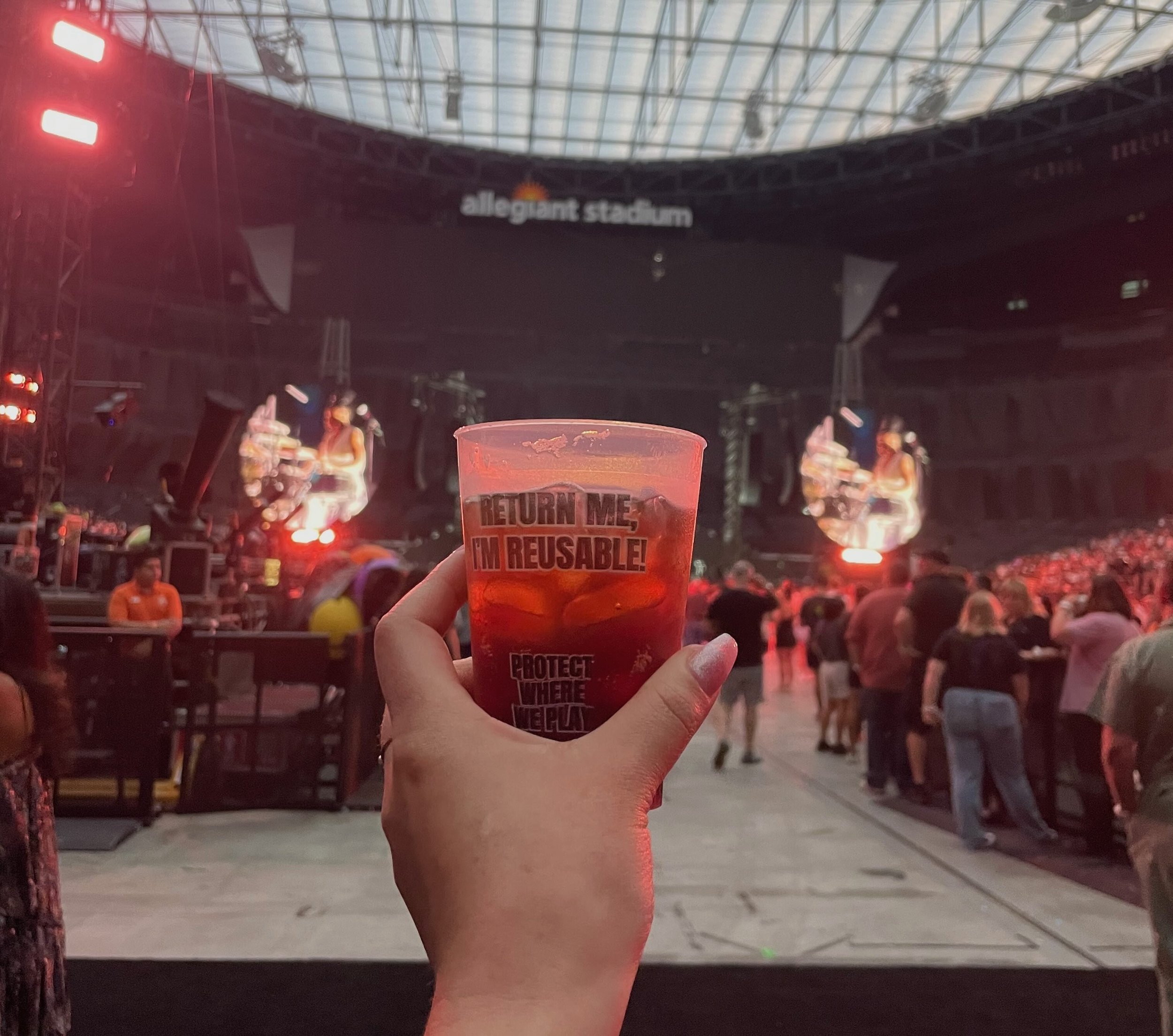 First-person POV shot of a hand holding a reusable cup at the Protect Where We Play tour, reading 'RETURN ME, I'M REUSABLE!'