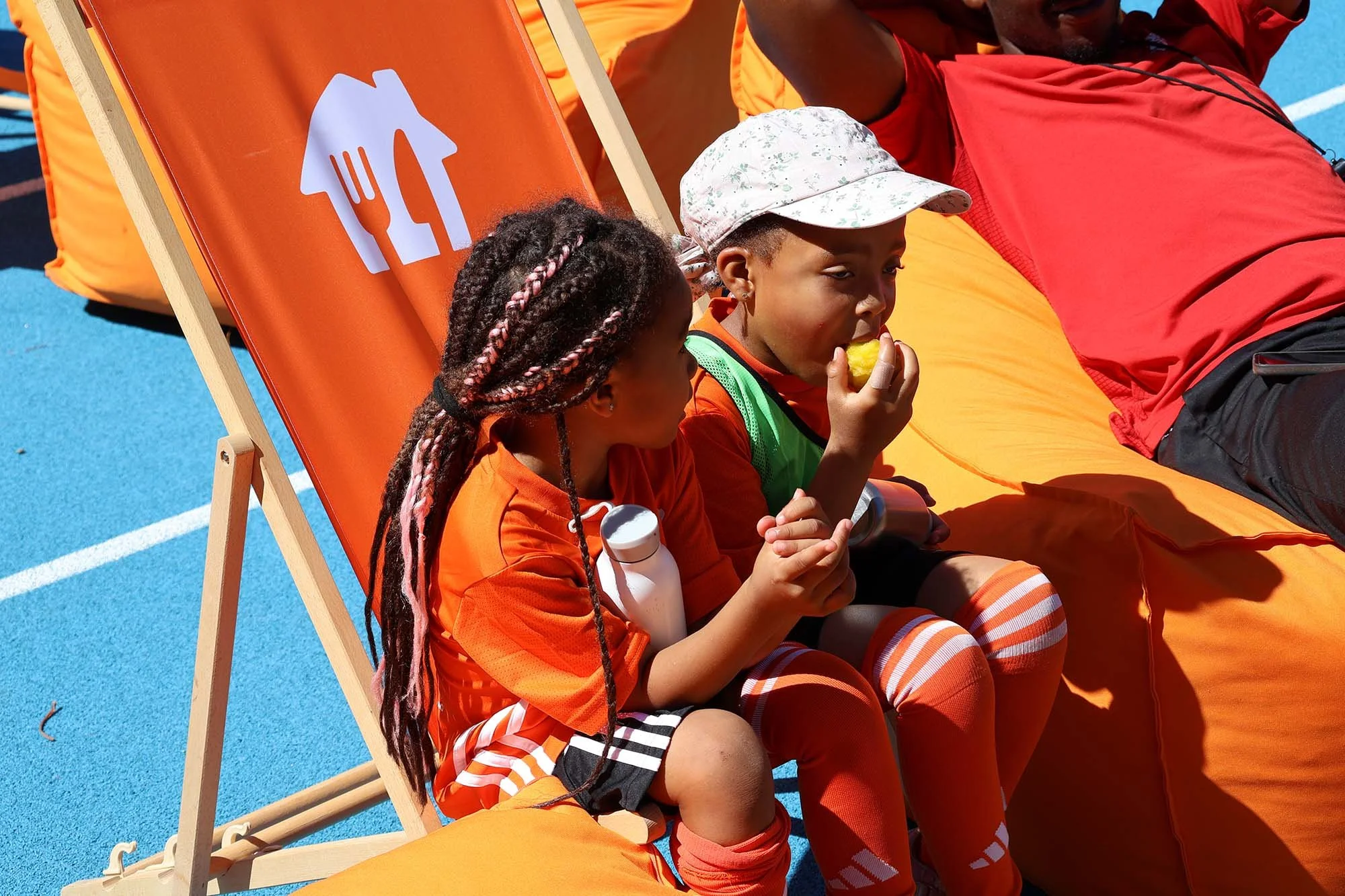 Two children sit on a deck chair eating together at a Feed the Game camp.
