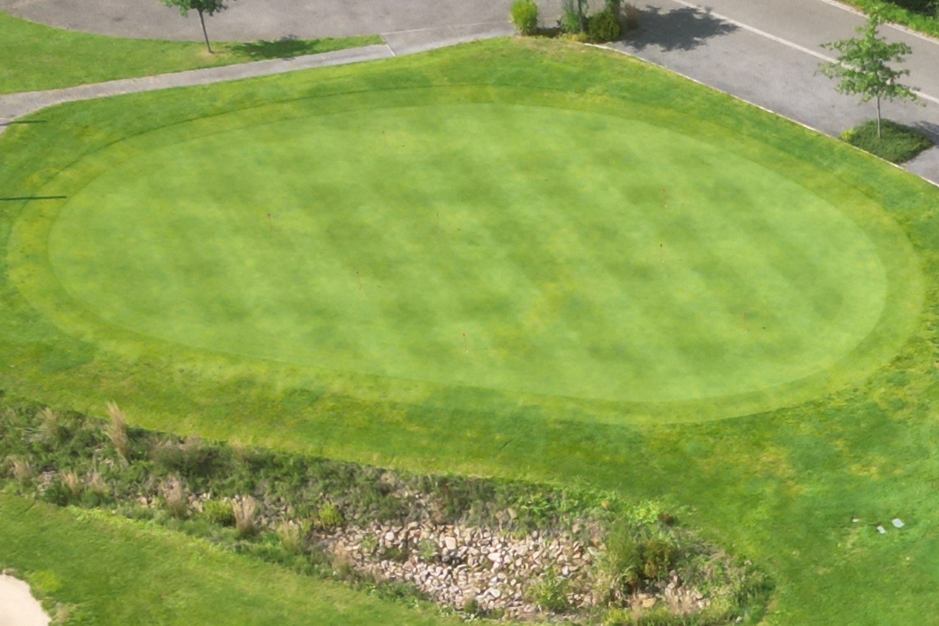Luftaufnahme eines gepflegten, ovalen Golfplatzes mit angrenzenden Wegen und Bäumen.