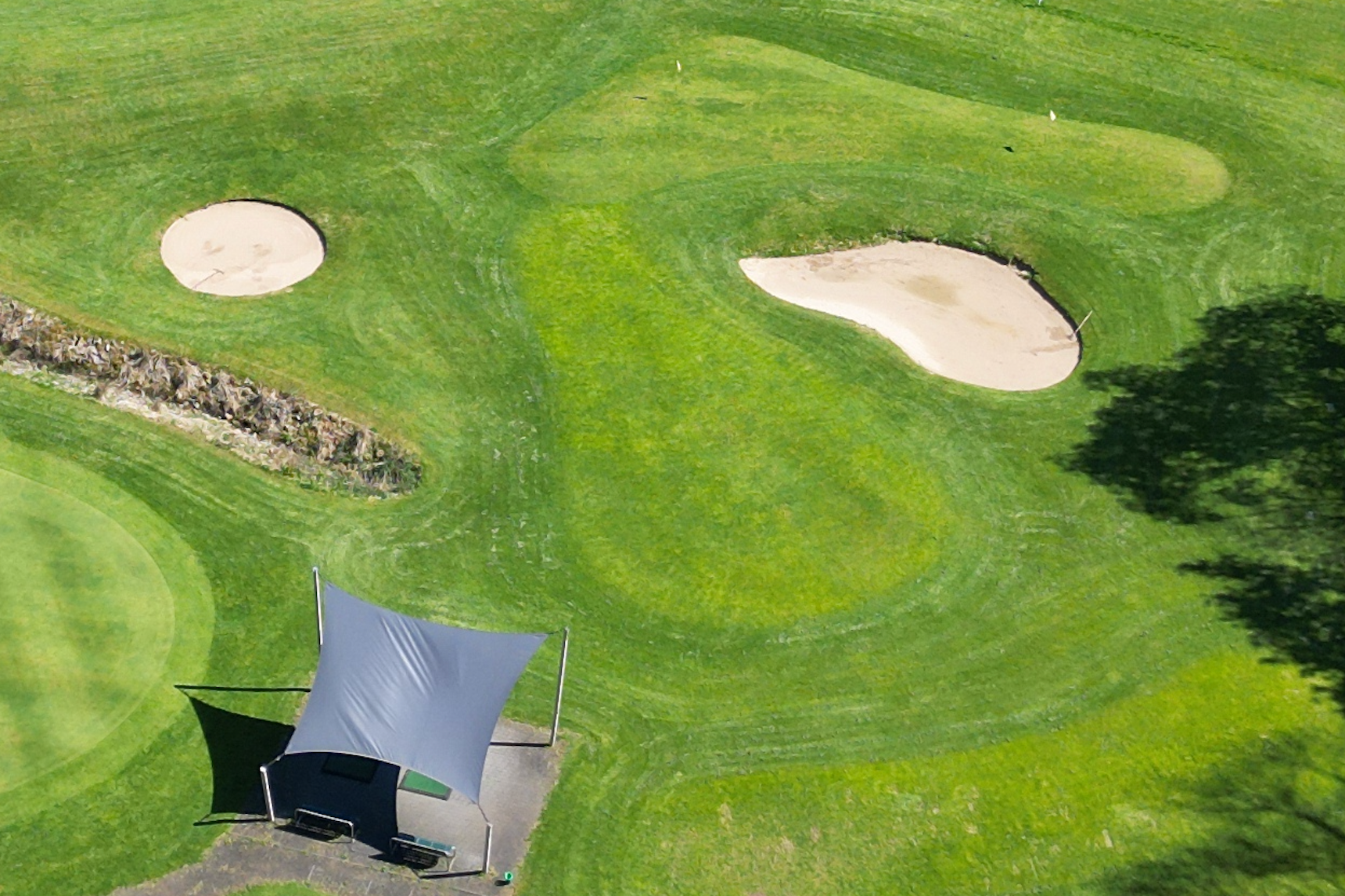 Luftaufnahme eines Golfplatzes mit zwei Bunkern, einem Wasserhindernis, einem Abschlag und einem Fairway, umgeben von Bäumen.
