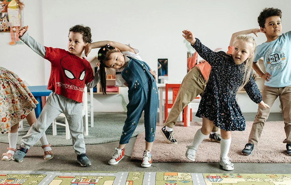 Children standing in a line with arms raised and leaning to the side in a classroom.