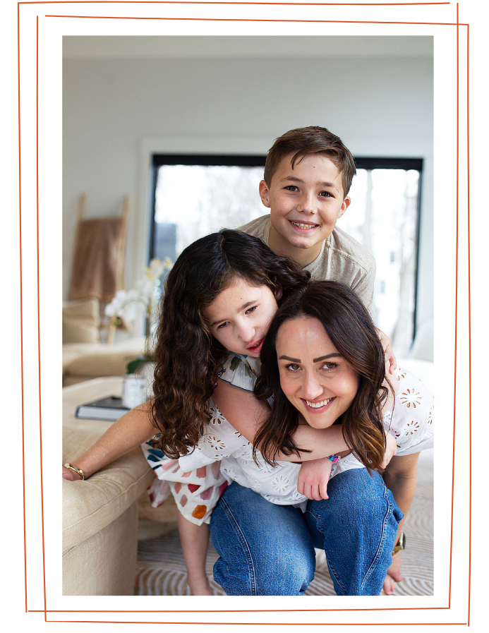 A woman with two children inside a home, with light-colored walls and large windows in the background. The woman is smiling and kneeling on the floor, with a girl and boy on her back, all smiling and appearing happy.