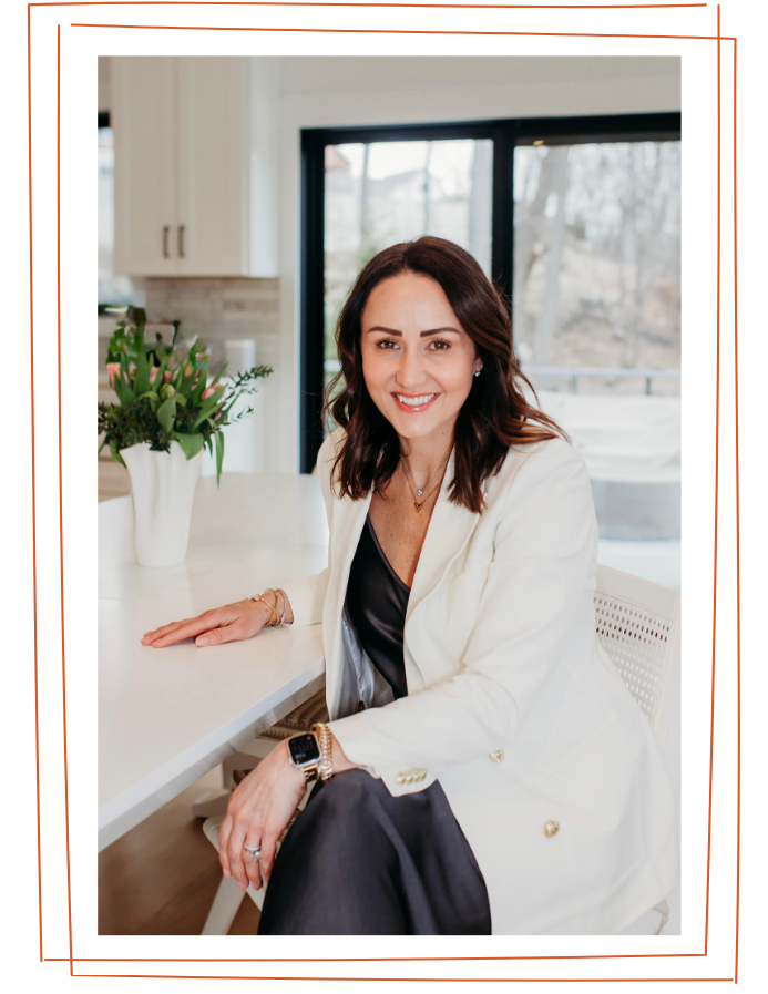 A woman with shoulder-length dark brown hair, wearing a white blazer, black dress, and accessorized with jewelry, sitting at a white kitchen counter with a potted plant, smiling at the camera.