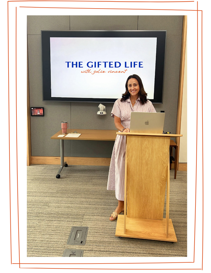 A woman standing at a wooden podium in front of a screen that displays the title "THE GIFTED LIFE with julie vincent." She is smiling, wearing a pink and white striped dress, and has a laptop in front of her. There is a table with a notebook and pink tumbler to her left, and the room has gray walls and a carpeted floor.