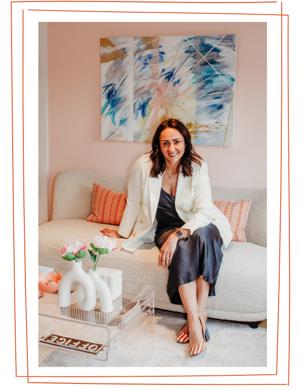 A woman with shoulder-length brown hair sitting on a beige couch with orange and beige striped pillows. She is smiling and wearing a white blazer over a dark dress, with striped orange and white shoes. Behind her is a colorful abstract painting on a pink wall. On the coffee table in front of her, there is a white decorative vase with pink and white flowers, a small orange bowl, and the word "DIFFERENT" spelled out in black letters on a white background.