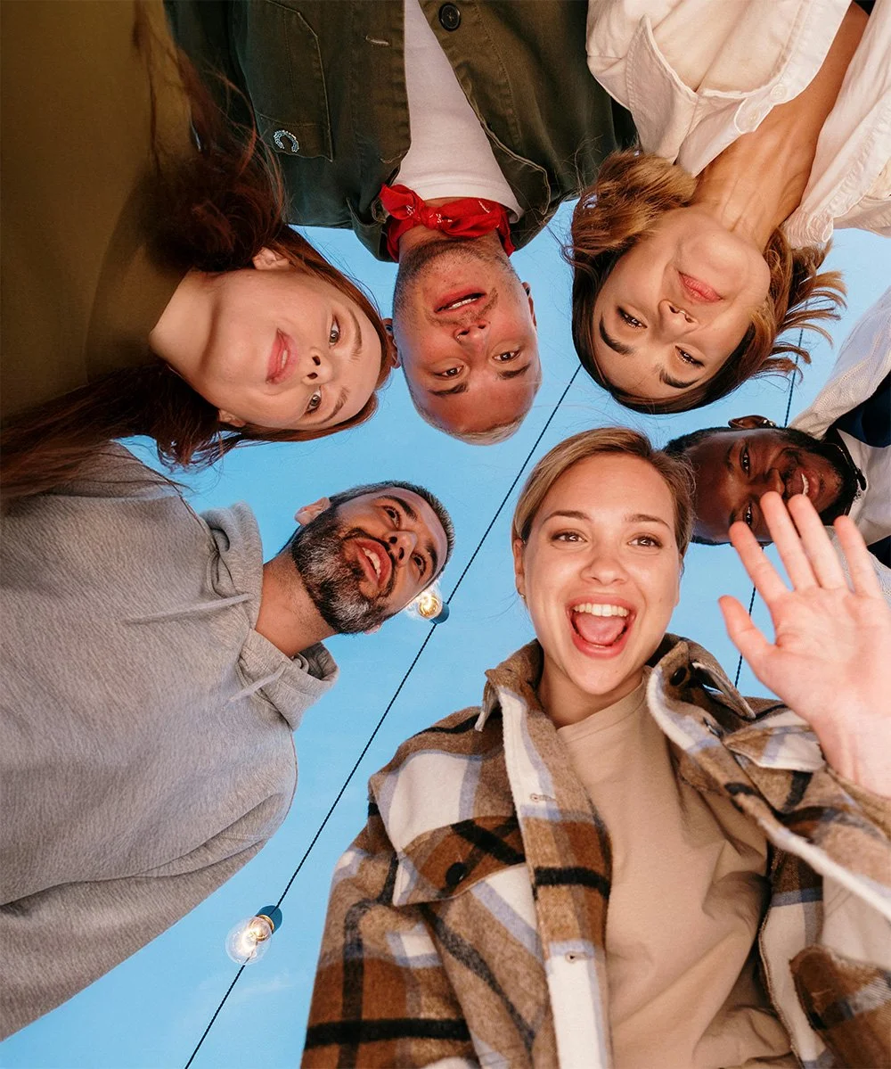Group of six diverse friends in a circle, looking down at the camera, smiling and waving, outdoors with string lights and blue sky in the background.