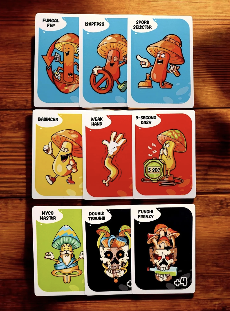 A set of illustrated game cards with mushroom characters displaying various traits and actions, including fungal flip, leapfrog, spore selector, balancer, weak hand, 5-second dash, myco master, double trouble, and funghi frenzy, laid out on a wooden 