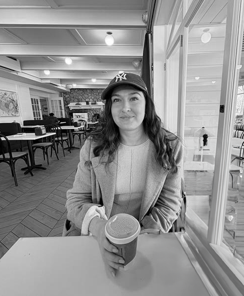 A woman sitting at a table in a cafe, holding a coffee cup, wearing a baseball cap and a coat, with a cozy indoor setting background.