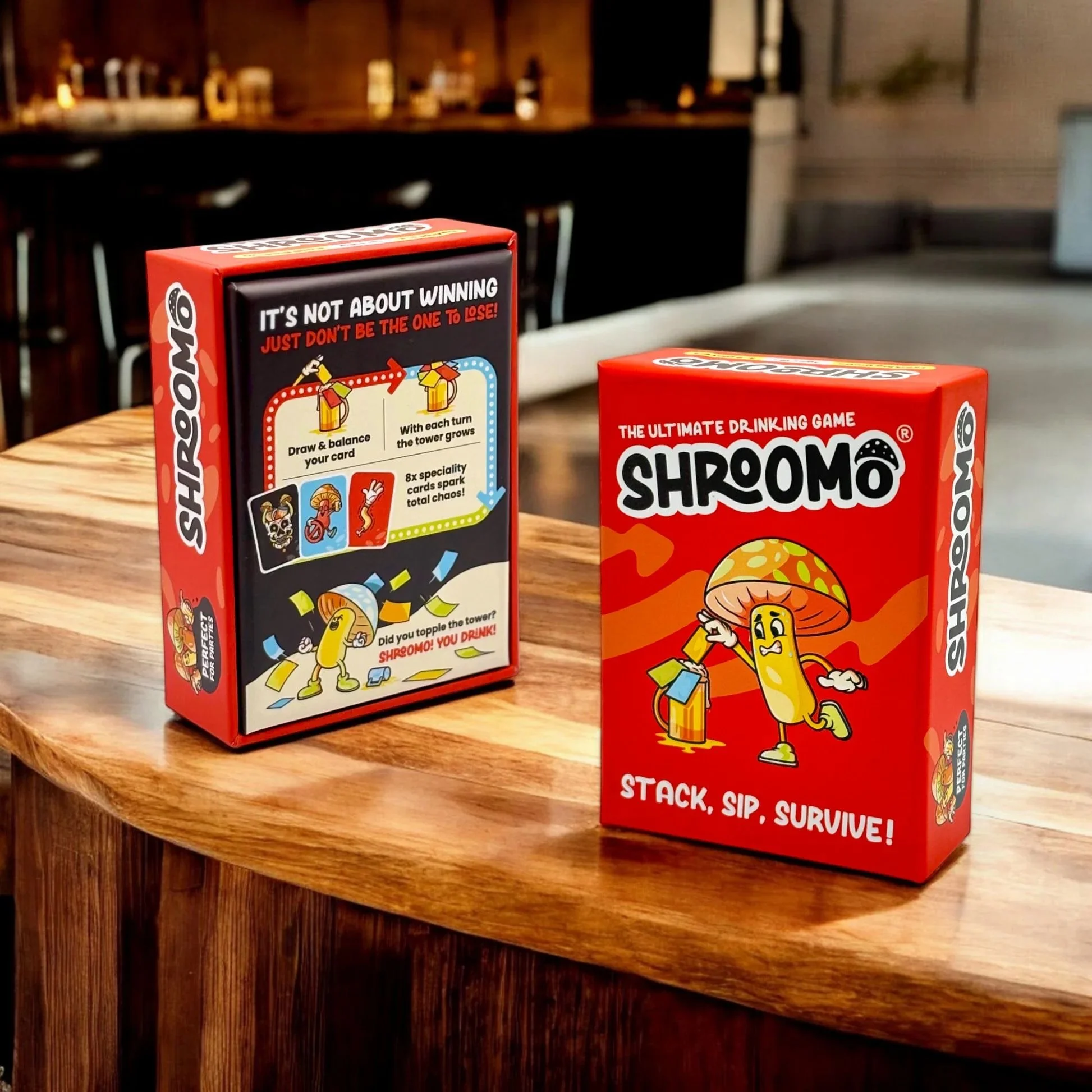 Boxes of Shromo, a drinking game, on a wooden table, one showing game instructions and the other with a cartoon mushroom character holding a beer.