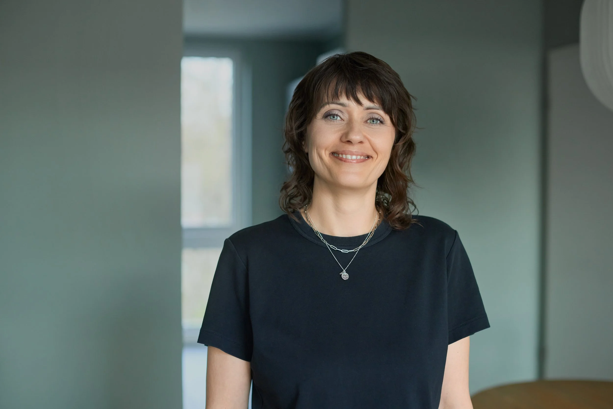 Nadine Dunkel. Sie trägt ein schwarzes T-Shirt und steht freundlich lachend in einem hellen Raum mit Fenster im Hintergrund.