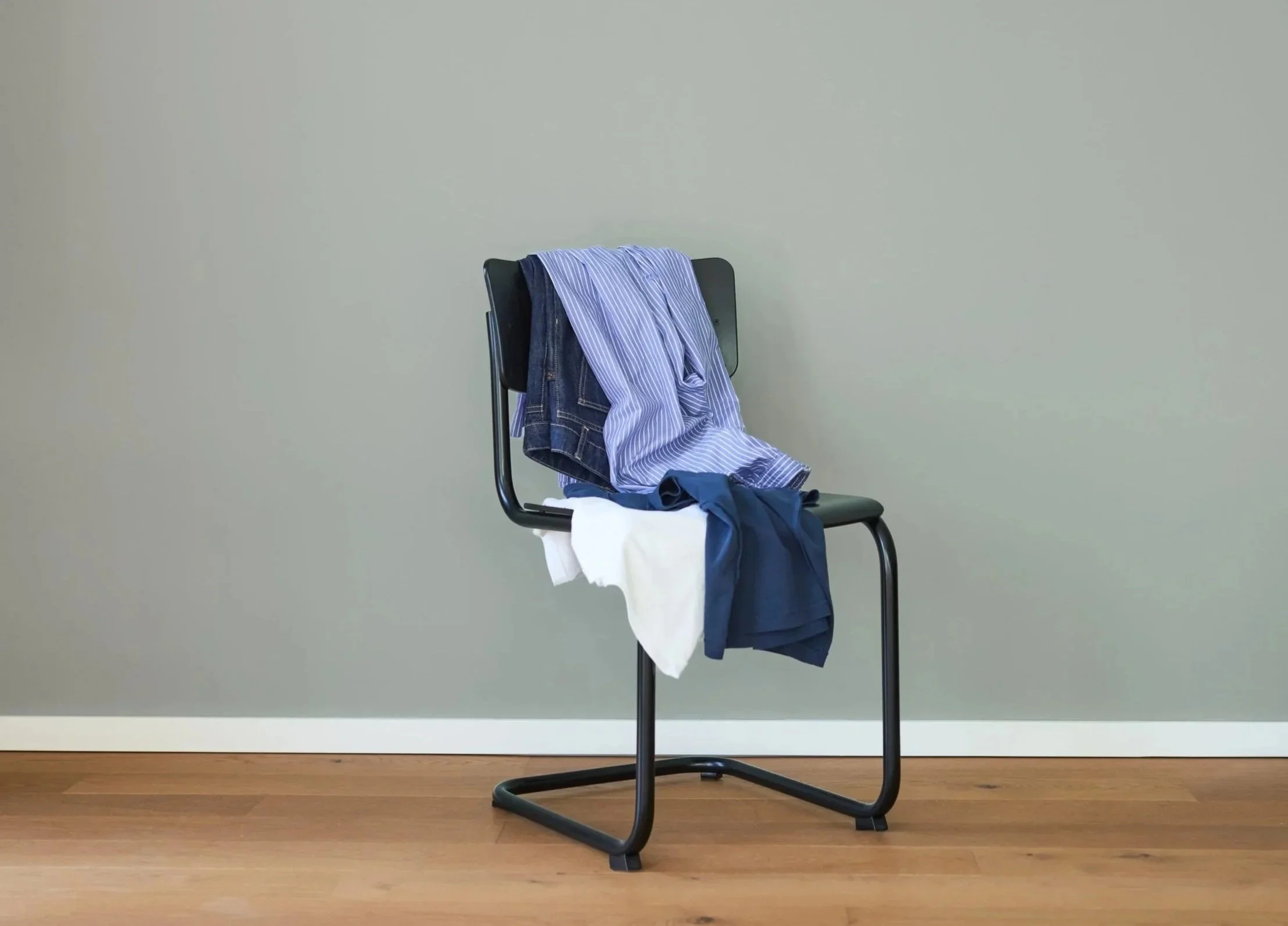 Ein schwarzer Stuhl mit Kleidung darauf, bestehend aus mehreren Hosen und T-Shirts. Der Stuhl steht vor einer leeren Wand auf einem Holzboden.