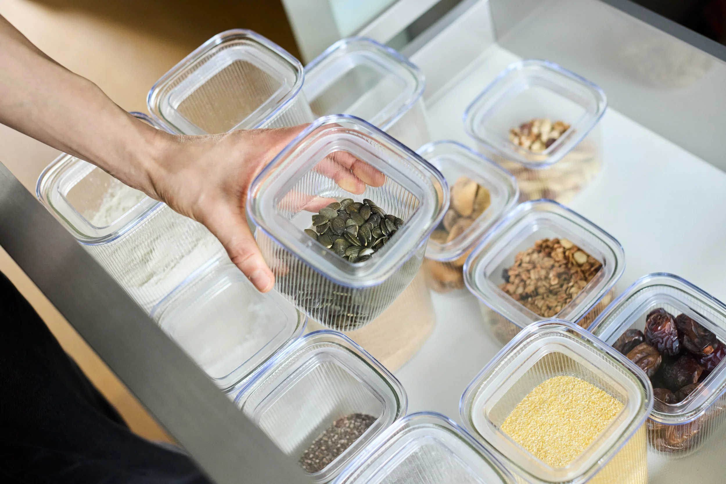 Eine Hand hält eine transparente Vorratsdose mit Kürbiskernen, umgeben von weiteren Vorratsgläsern mit verschiedenen Lebensmitteln in einer Küchenschublade.
