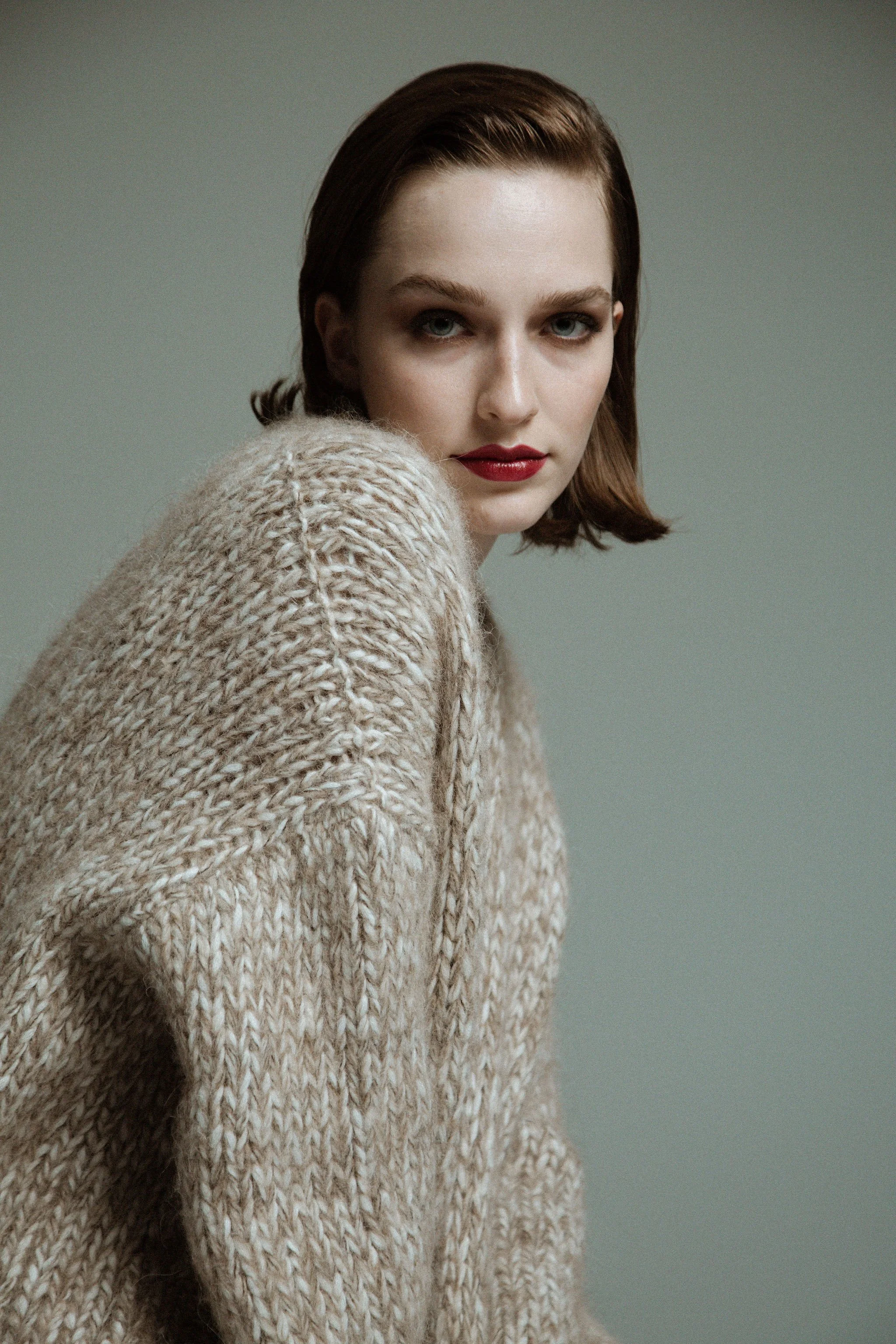 A woman with short brown hair wearing a chunky beige knit sweater, looking directly at the camera with a neutral expression.