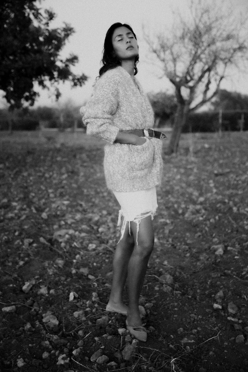 A woman stands outdoors on rocky terrain with bare trees in the background, wearing a sweater, ripped shorts, and high heels, with one hand in her pocket and eyes closed.
