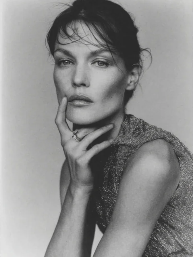 Black and white portrait of a woman with short hair, resting her chin on her hand, wearing a sleeveless top with a textured pattern.