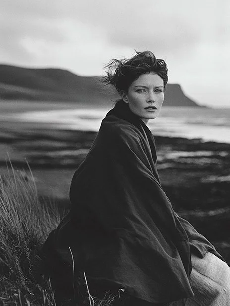 A woman sitting on grass near the coast, with ocean waves and cliffs in the background, photographed in black and white. Ai Generated, Creative direction with  AI 