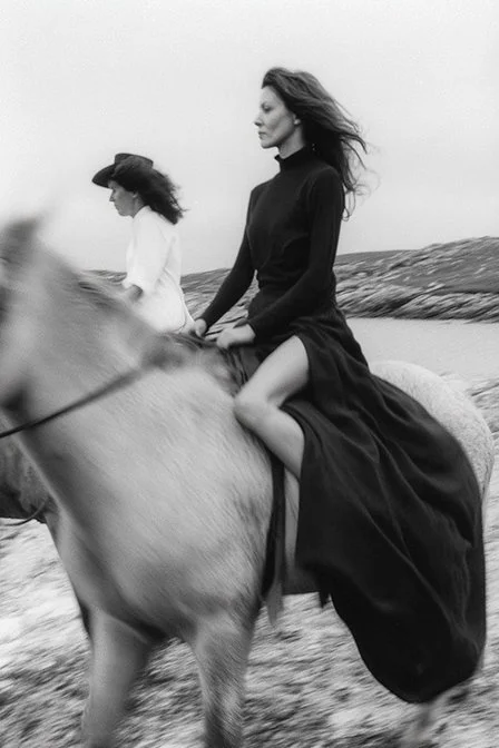 A woman riding a horse along a beach with another woman walking nearby, both wearing black and white clothing. Ai Generated, Creative direction with  AI 