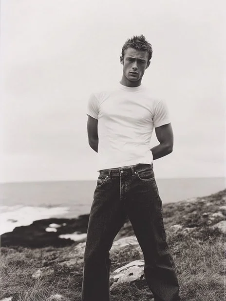 A young man standing outdoors on a rocky terrain near the ocean, wearing a white t-shirt and dark jeans, with his hands behind his back, looking at the camera. Ai Generated, Creative direction with  AI 