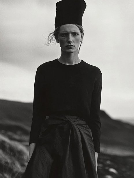 A woman standing outdoors on a cloudy day, wearing a long-sleeved black top with a tied jacket around her waist and a tall black hat, with a mountainous landscape in the background. Ai Generated, Creative direction with  AI 