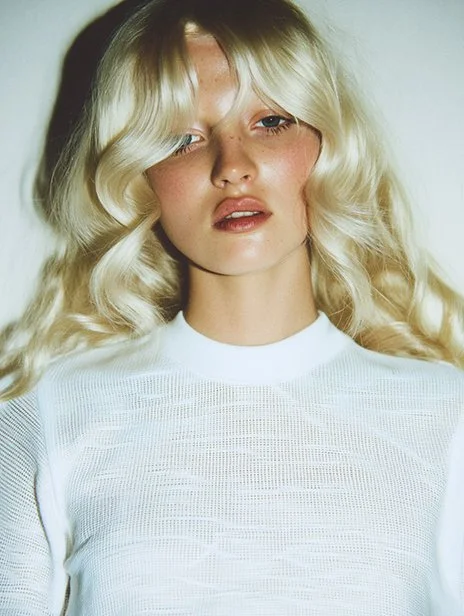 A young woman with long, wavy blonde hair and fair skin wearing a white top with a textured pattern, looking slightly to the side with a neutral expression. Ai Generated, Creative direction with  AI 