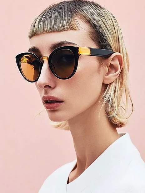 A young woman with short blonde hair wearing large black sunglasses with gold accents and a white top, against a pink background. Ai Generated, Creative direction with  AI 