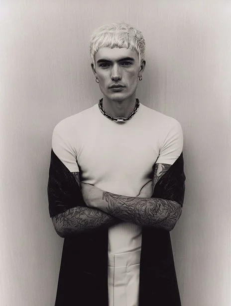 Black and white photo of a young man with short, bleached hair, wearing a white t-shirt, a chunky chain necklace, and tattoos on his arms, standing against a plain wall. Ai Generated, Creative direction with  AI 