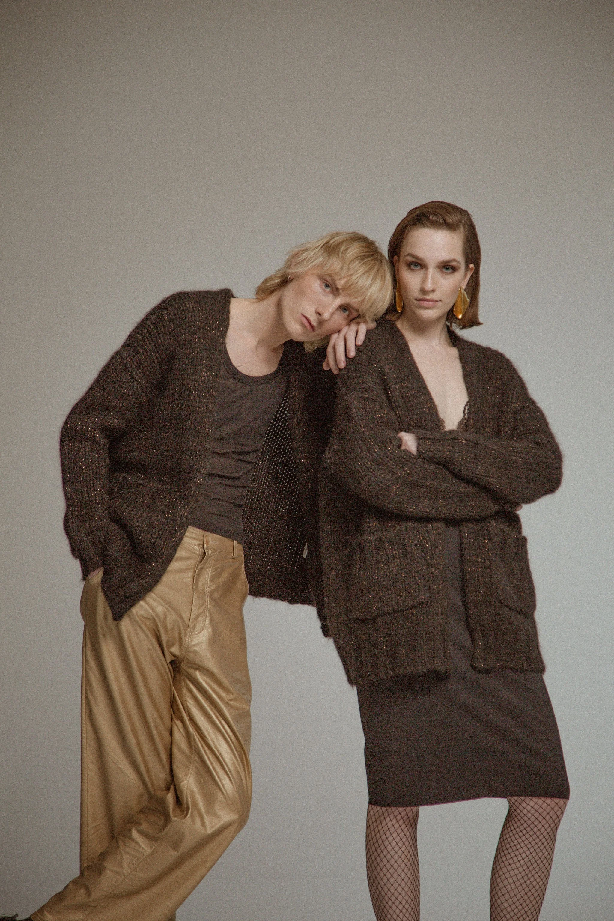 Two women standing close together against a neutral background, dressed in dark cardigans and skirts, with one woman resting her head on the other's shoulder.