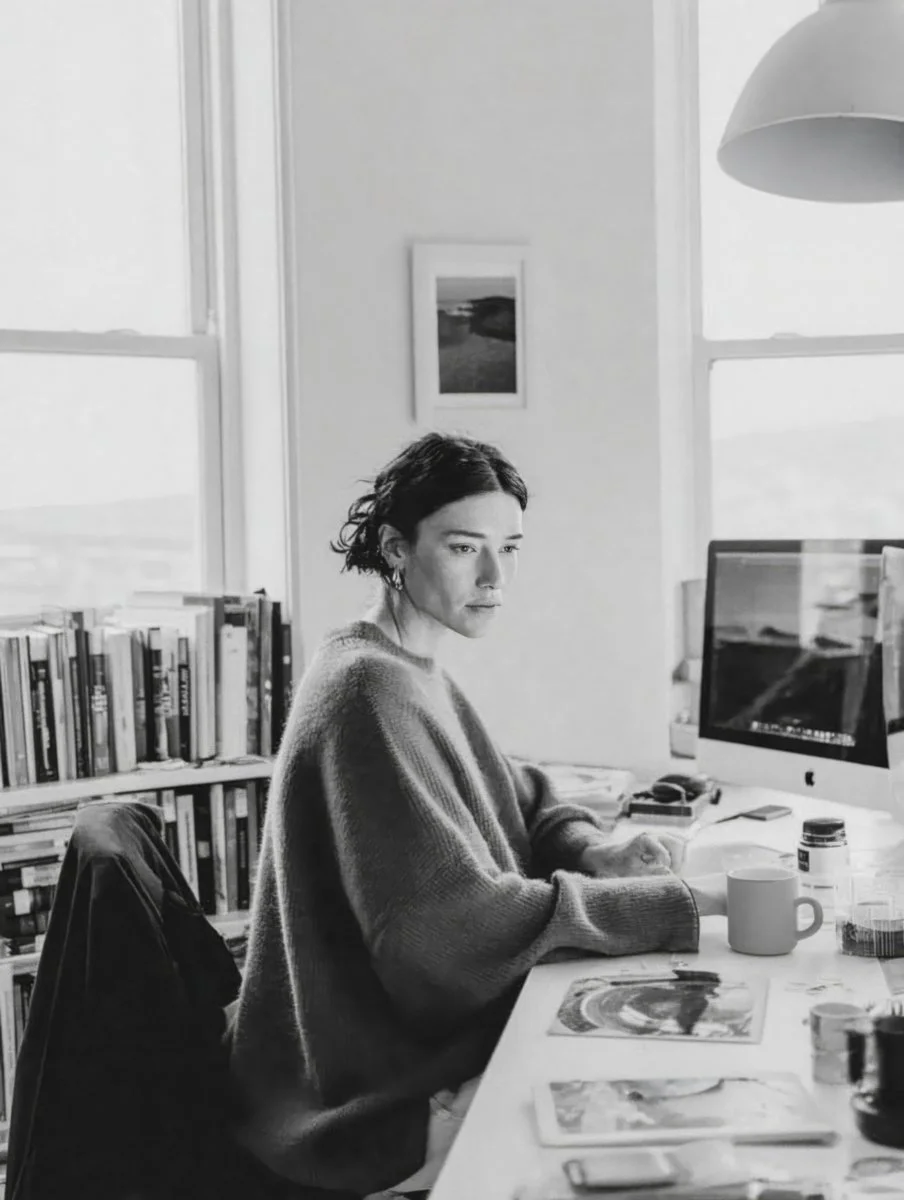A woman with short curly hair and a sweater sits at a desk in front of an iMac computer, surrounded by books, papers, and art supplies in a bright room with large window windows.