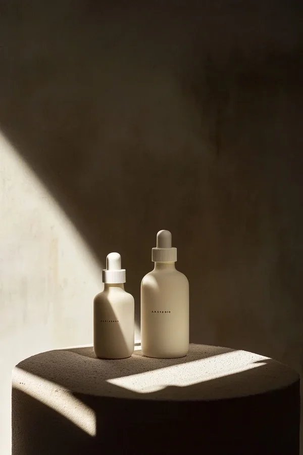 Two minimalist skincare bottles with dropper caps on a rounded beige pedestal, lit by natural sunlight creating shadows, against a textured wall background.