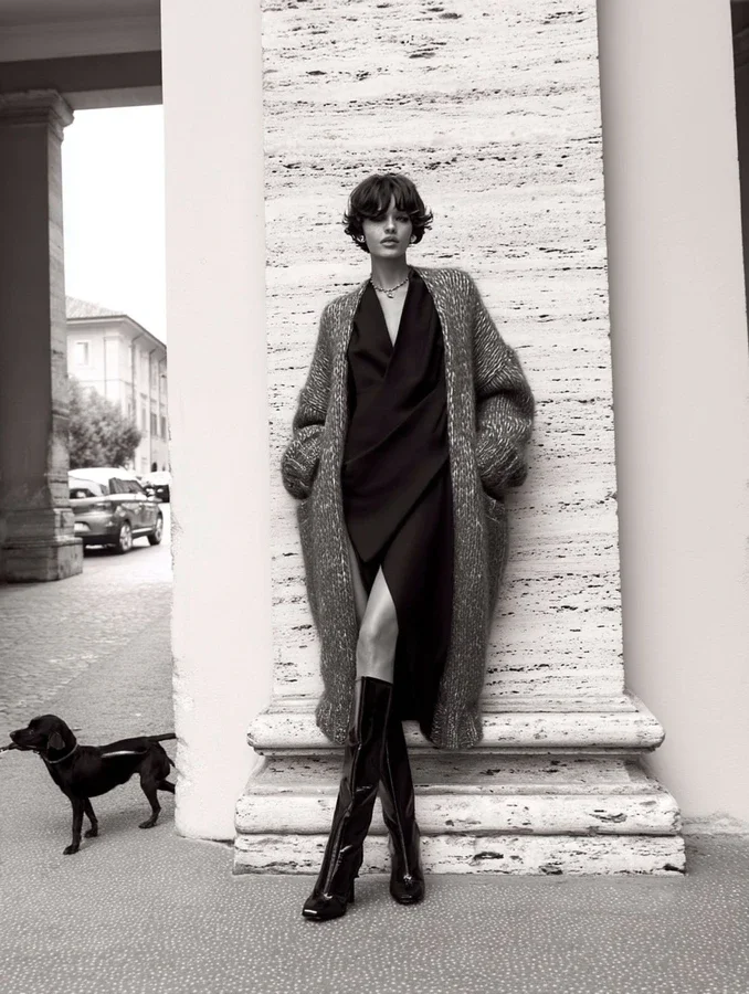 A woman with short wavy hair standing against a textured stone wall, wearing a long coat, dress, and knee-high boots. She is outdoors on a city street with parked cars and a building in the background, and a small black dog on a leash nearby.